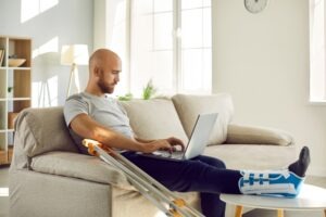 A man with a leg brace and crutches is sitting on a couch holding a laptop computer.