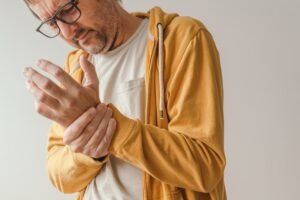 A middle-aged man in a white t-shirt and yellow hoodie is grasping his wrist.