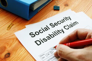 A person's hand holding a red pen poised to fill out a form that reads "Social Security Disability Claim."